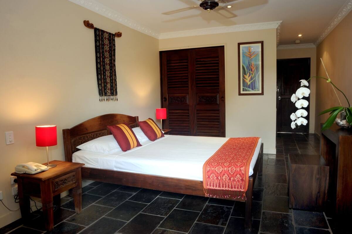 A hotel room with a wooden bed, two bedside lamps, a telephone, a red throw blanket, a wooden wardrobe, a painting, and a potted orchid.