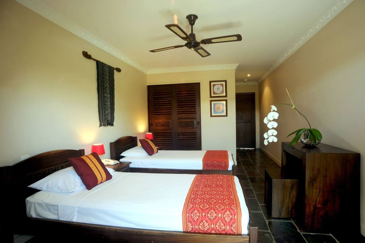 A hotel room with two beds, a ceiling fan, a wooden door, and a plant.