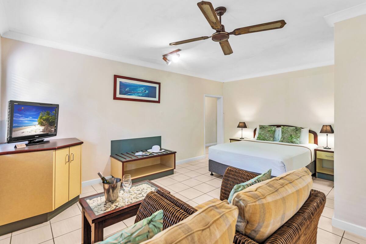 Une chambre d’hôtel avec un lit, une télévision, un ventilateur de plafond, une petite table avec une bouteille et des verres, et un tableau encadré au mur.