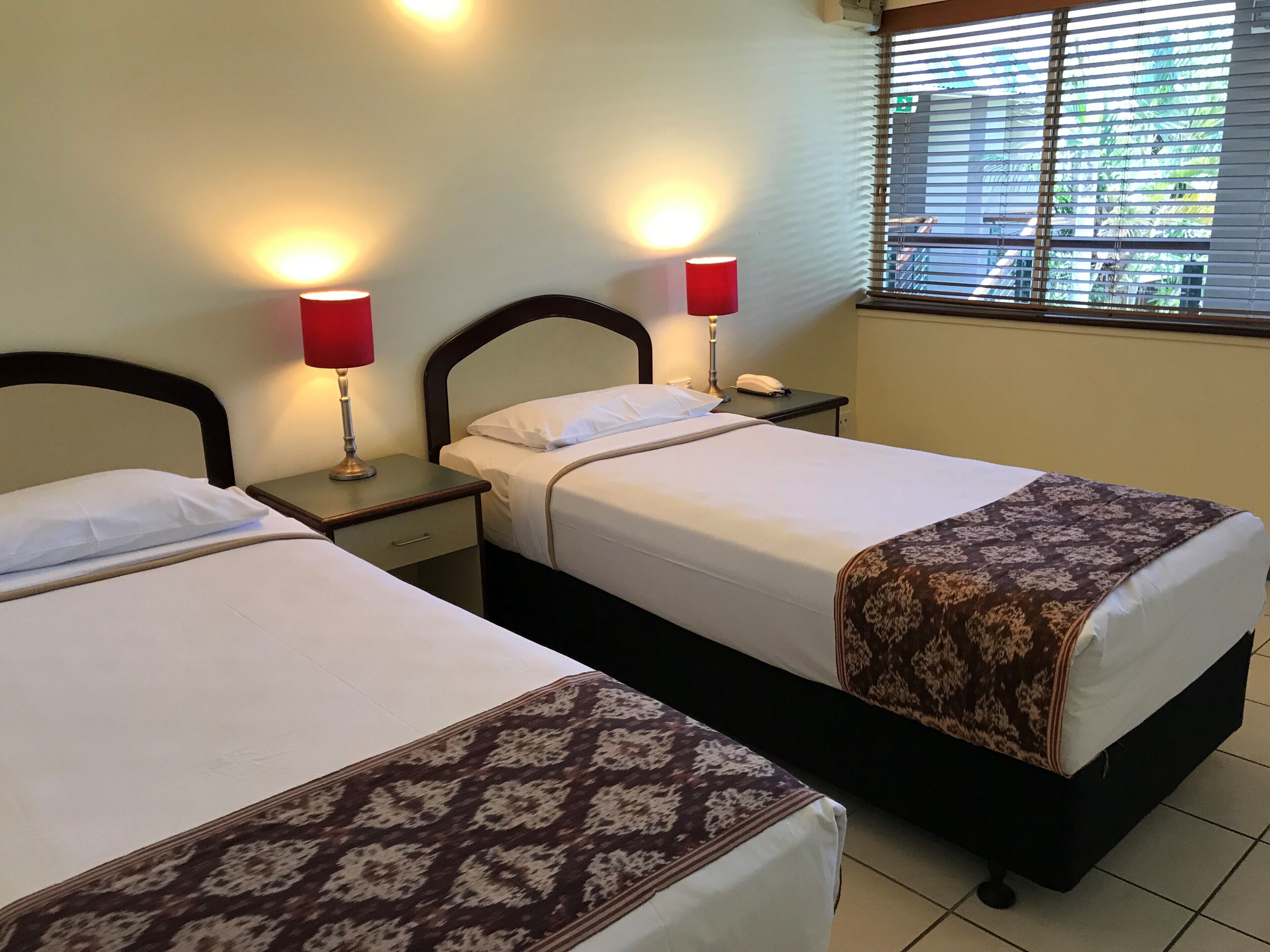 Two beds with headboards, bedside tables, lamps, and a window with blinds.