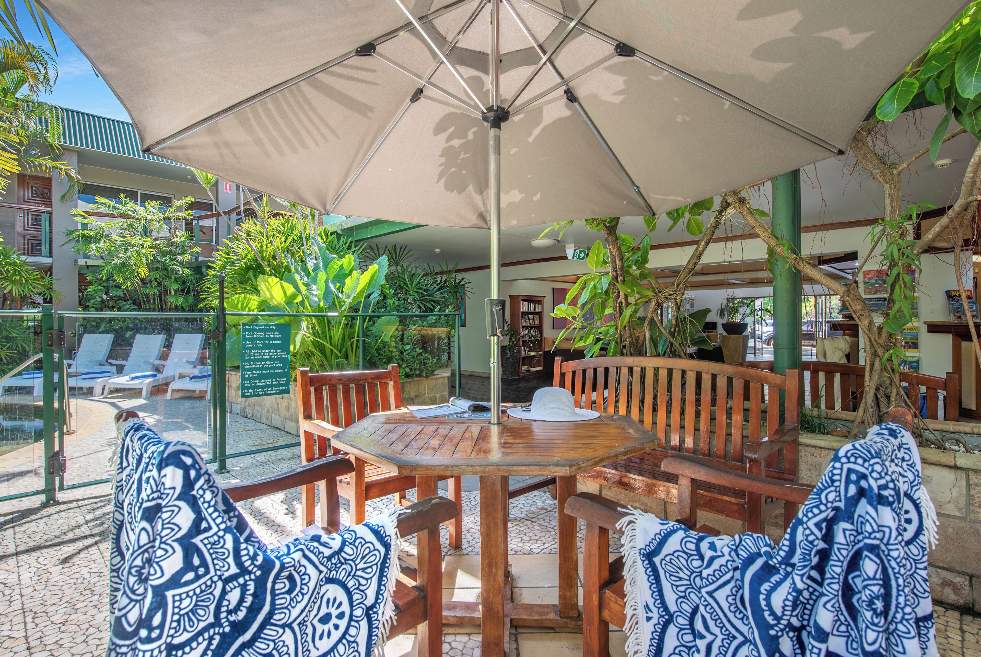 Uma área externa com espaço para sentar, uma mesa redonda, cadeiras de madeira e um grande guarda-sol.