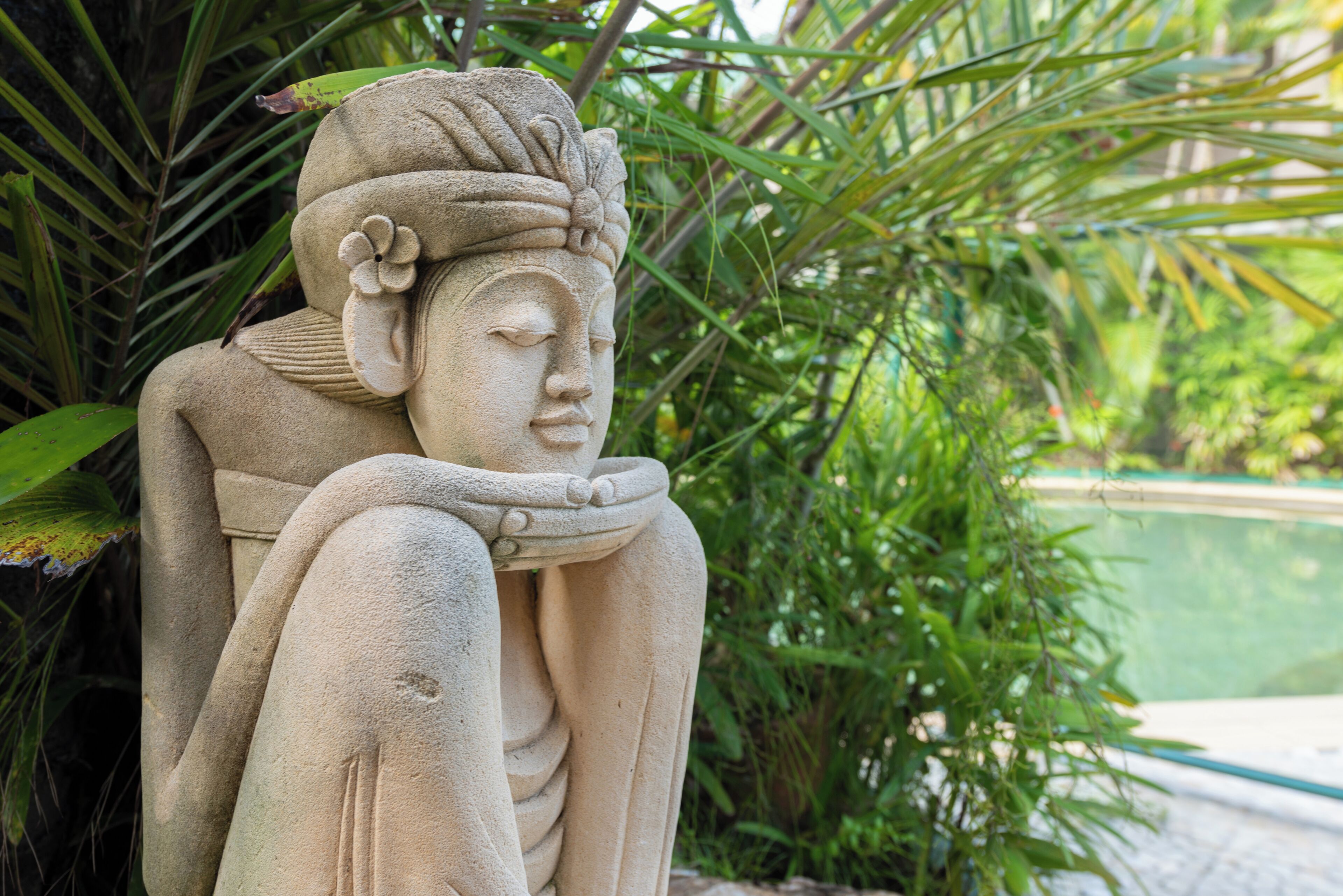 A stone statue of a seated figure with a flower in its hair, surrounded by lush greenery.