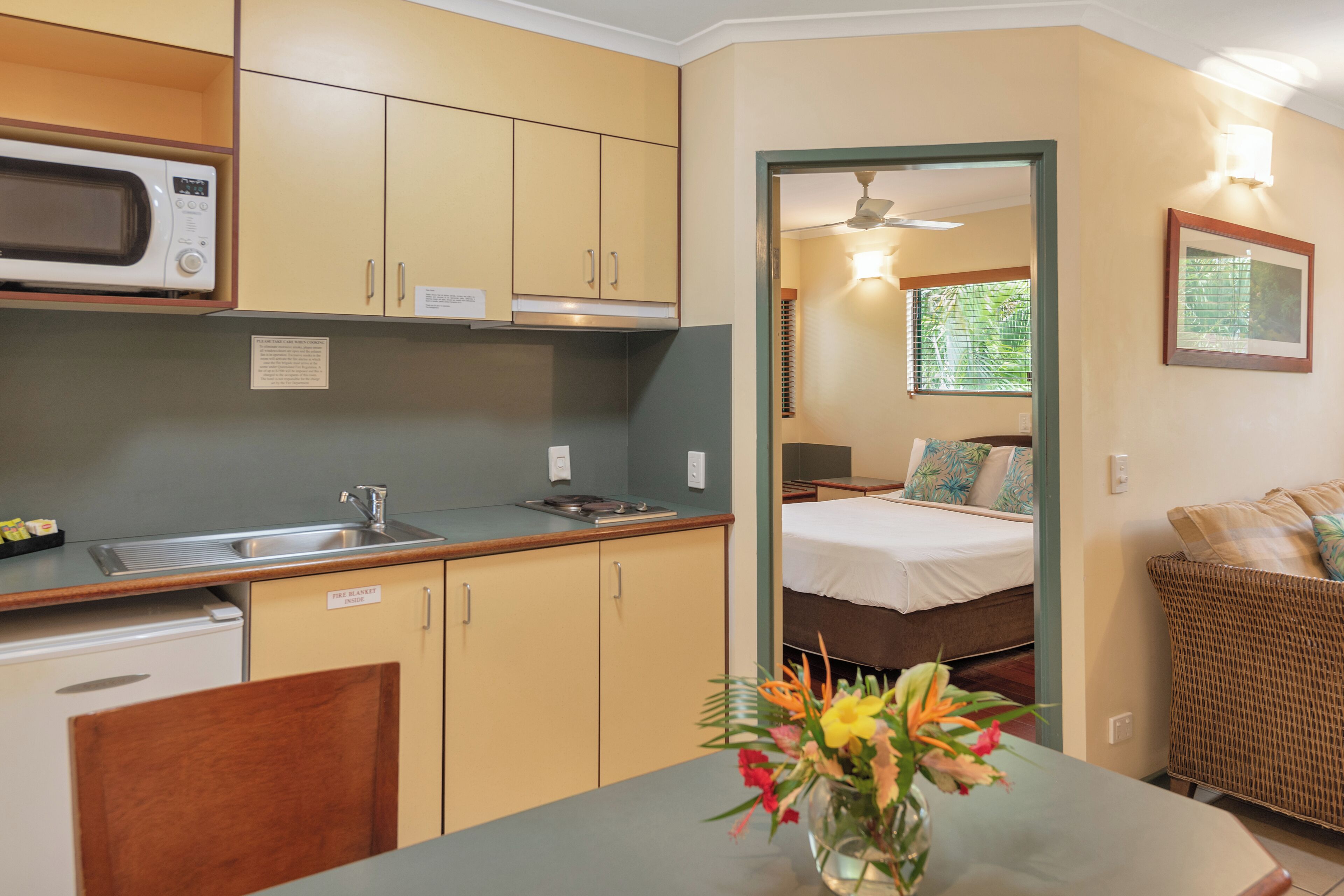 A compact hotel room with a kitchenette, a bed, a wicker chair, and a view of the outdoors.