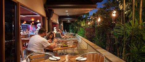 Un área de comedor al aire libre con mesas y sillas, rodeada de follaje y un paseo techado.