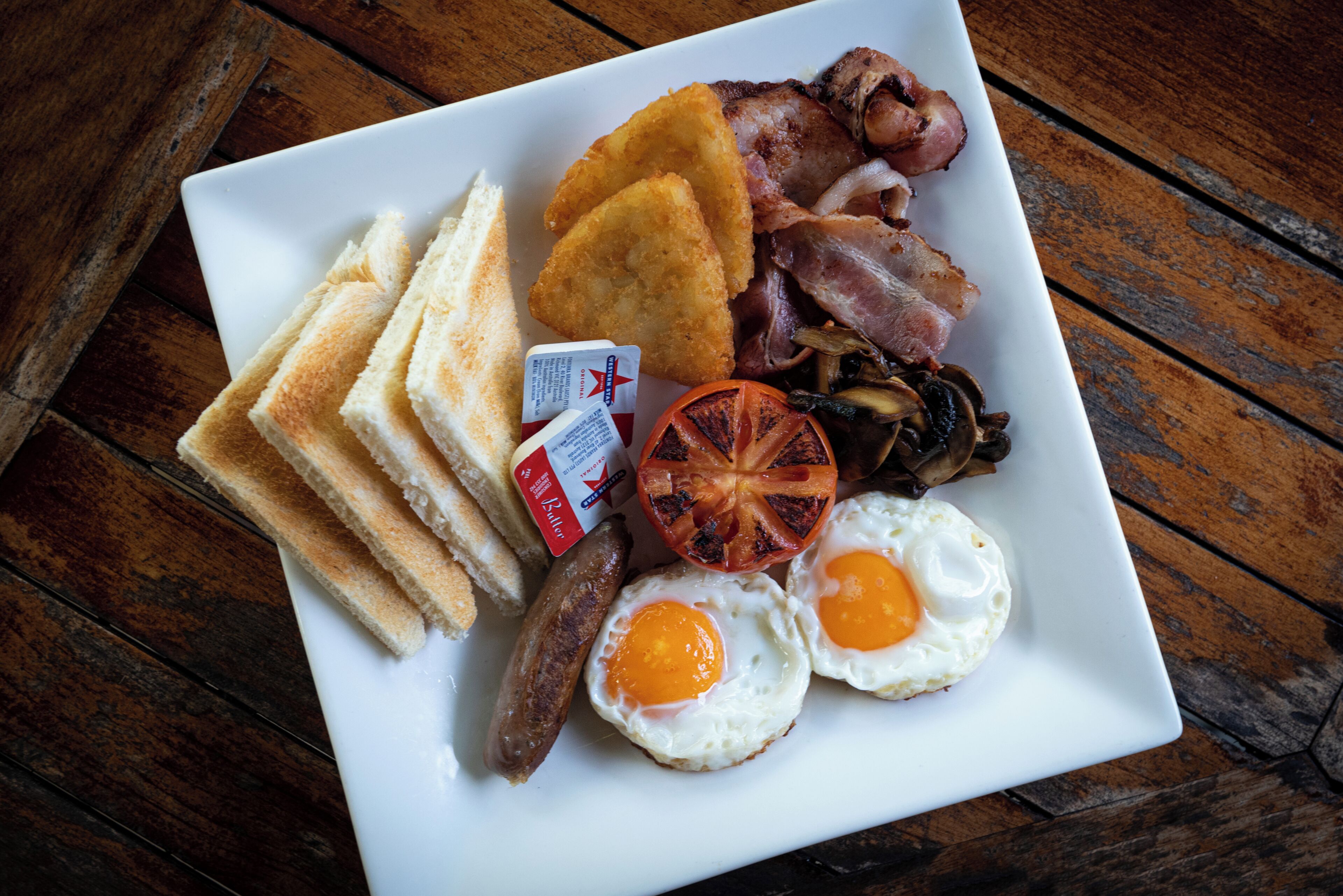 A plate of breakfast food including toast, sausages, eggs, bacon, and a grilled tomato.