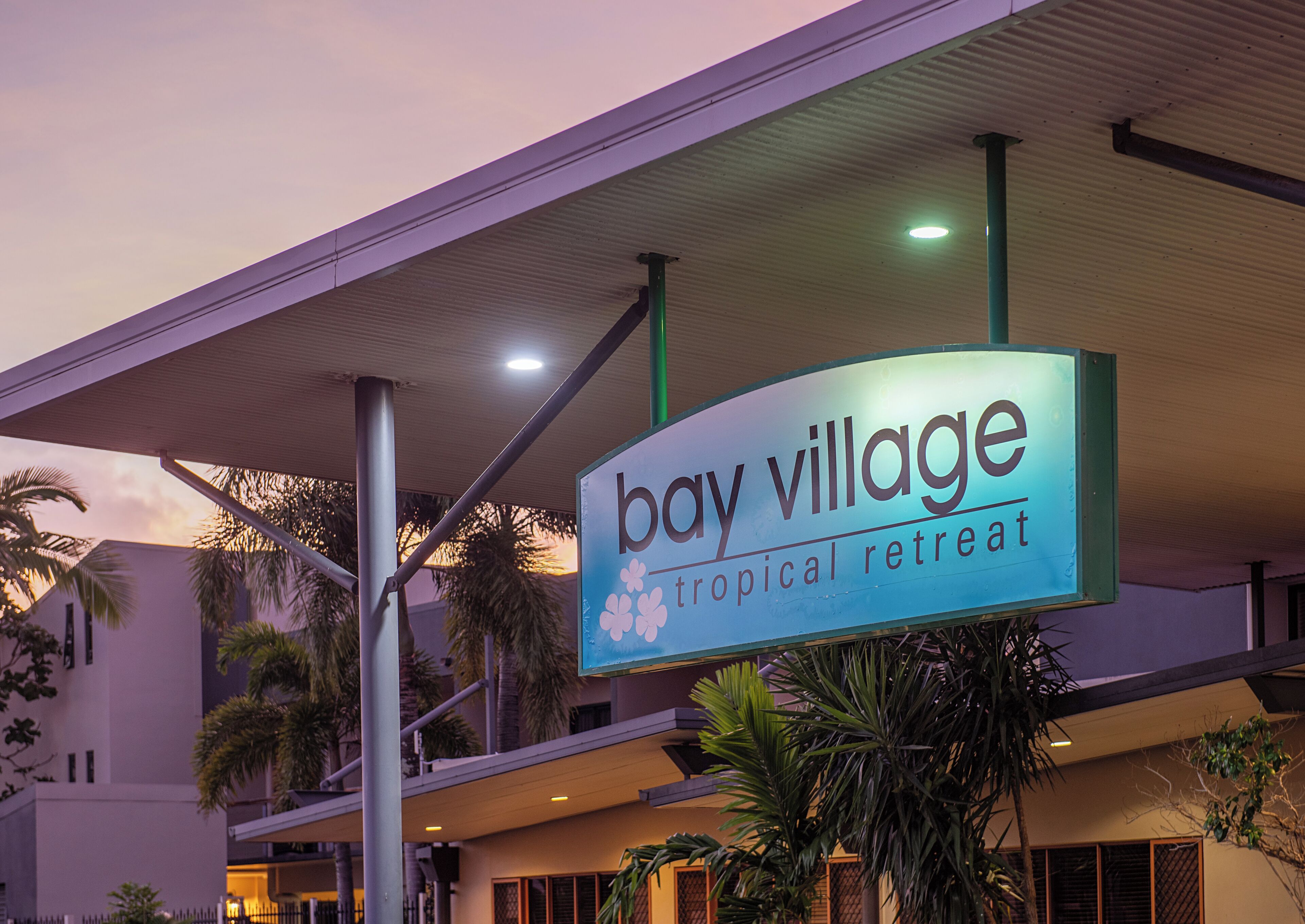 A sign for 'Bay Village Tropical Retreat' with a modern building and palm trees in the background.