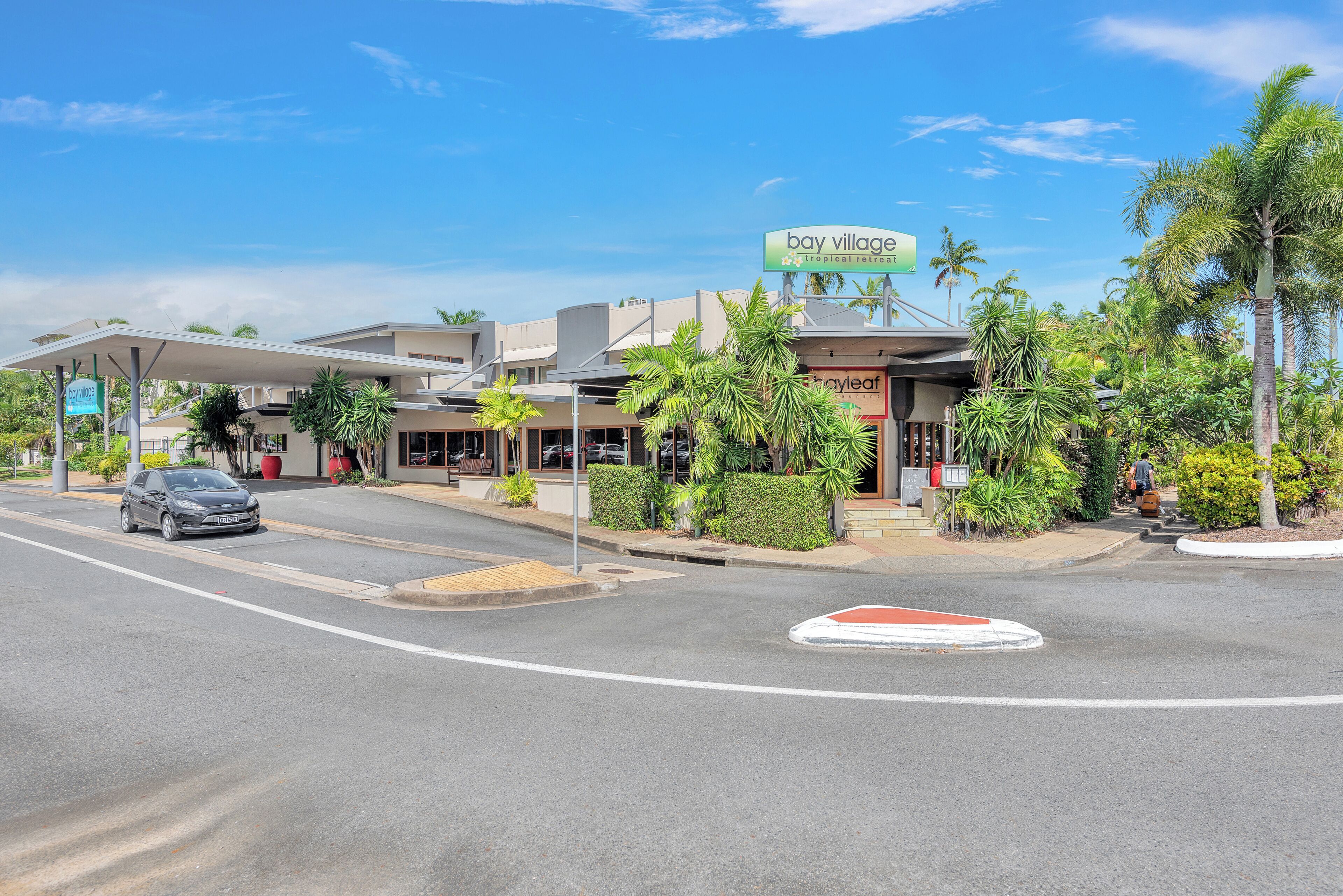A street view of a hotel named 'Bay Village' with a sign, palm trees, and a car parked on the road.