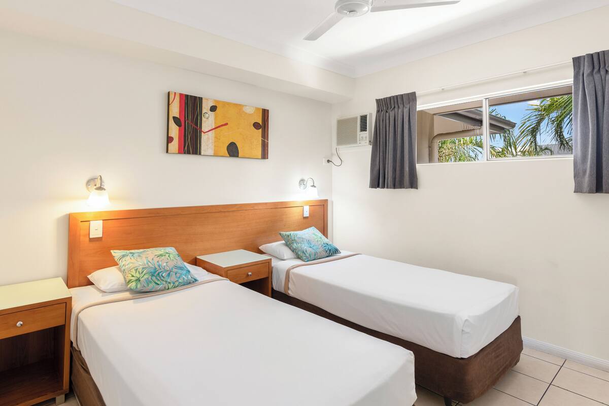 Une chambre d’hôtel avec deux lits simples, une tête de lit en bois, un ventilateur de plafond, une fenêtre avec des rideaux et un climatiseur fixé au mur.