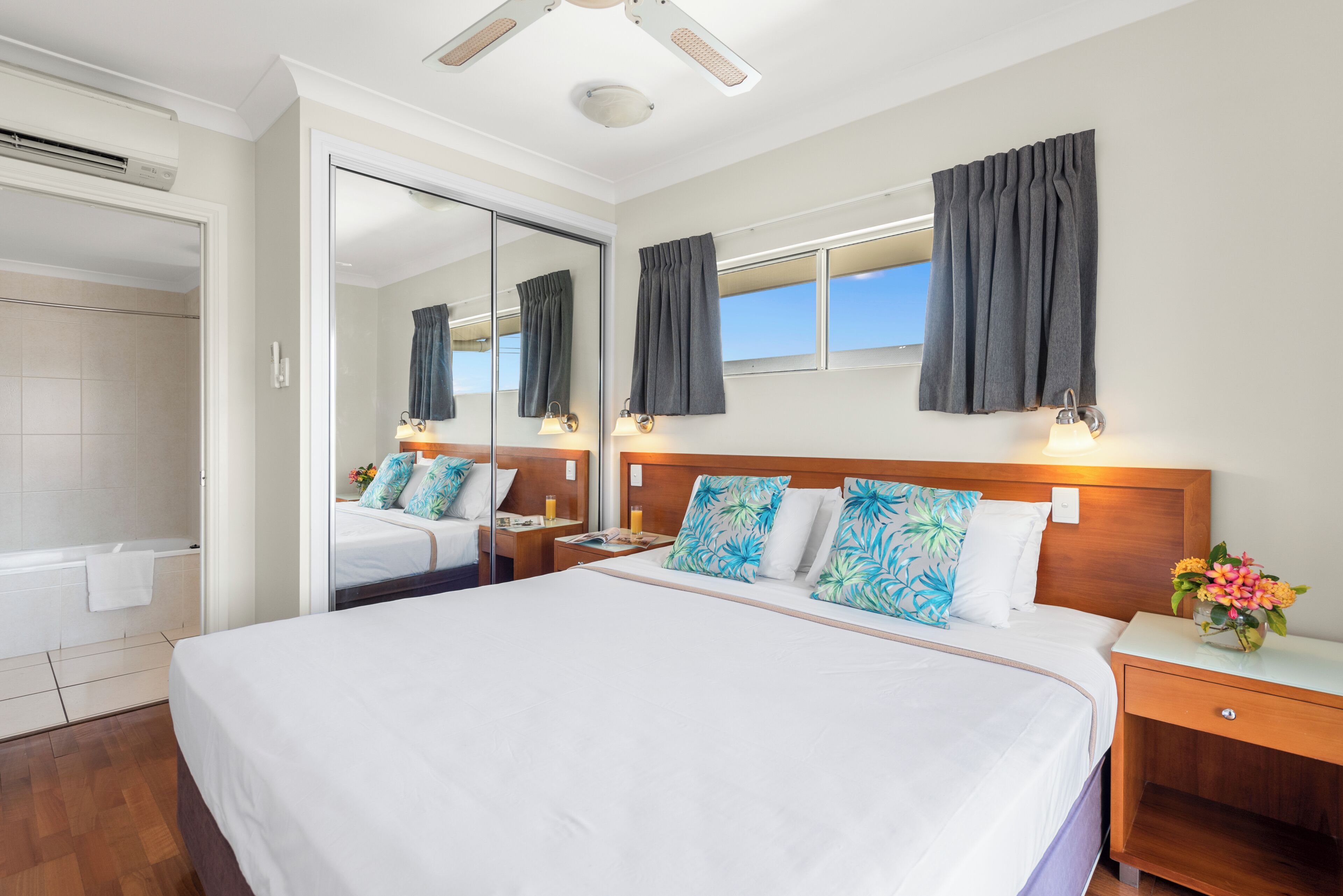 A hotel room with a large bed, a wooden headboard, a nightstand, a bedside table with a lamp, a mirror, a window with curtains, and a bathroom with a shower and bathtub.