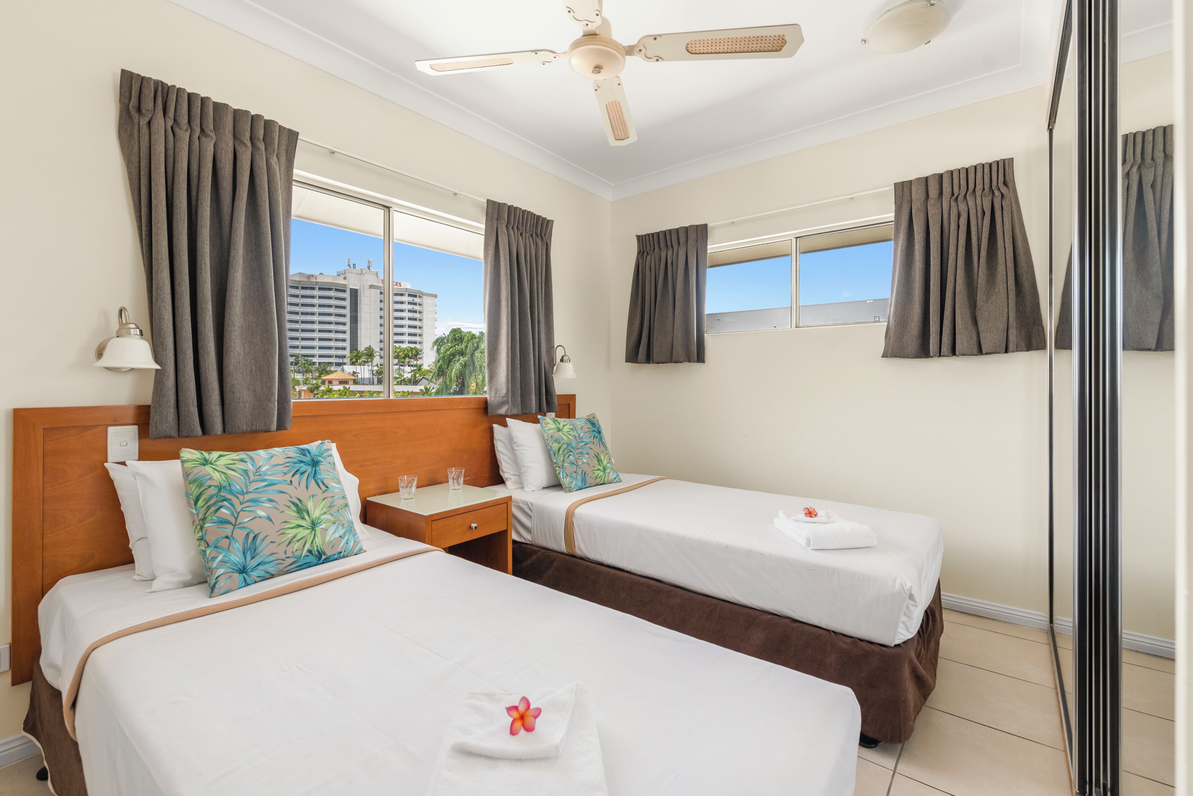 A hotel room with two beds, a ceiling fan, a window with curtains, and a view of buildings outside.