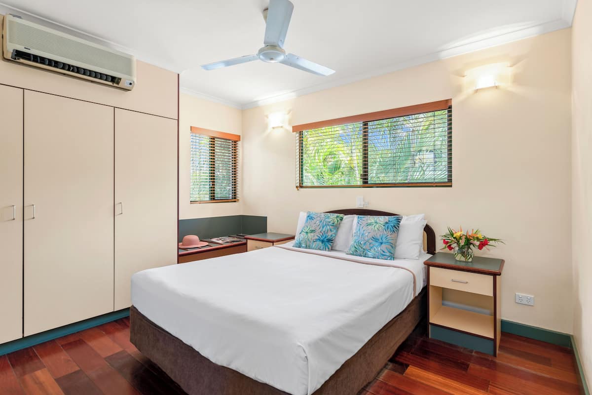 Une chambre à coucher comprenant un lit, des tables de chevet, une commode, un ventilateur de plafond, un appareil de climatisation et une fenêtre avec des stores.