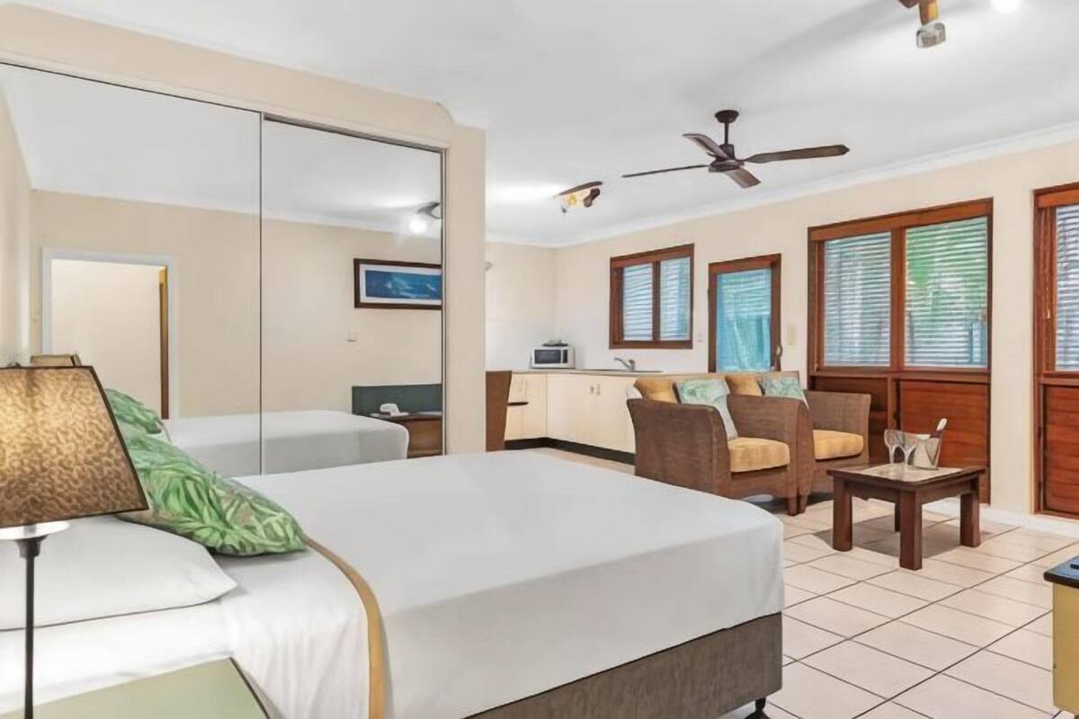 Une chambre d’hôtel comprenant un lit, un bureau, une chaise, une petite table, un ventilateur de plafond et une fenêtre avec des stores.