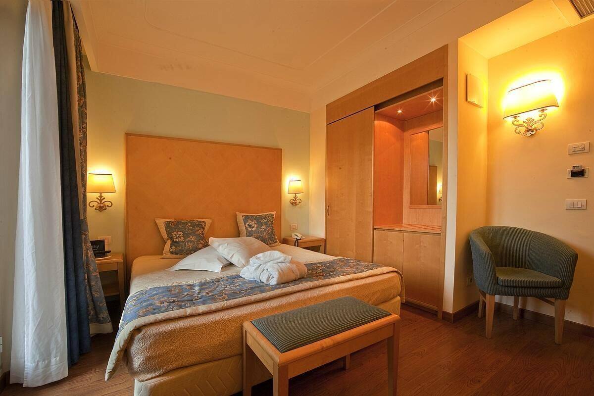 Una habitación de hotel con una cama, lámparas de noche, una silla, un banco y un espejo.