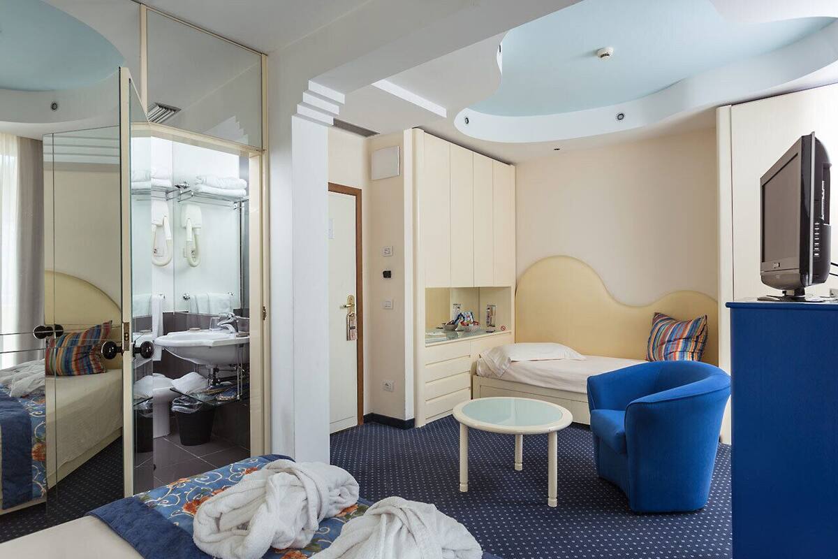 Habitación de hotel con cama, un sillón azul, una mesita, televisión y un baño con espejo y lavamanos.