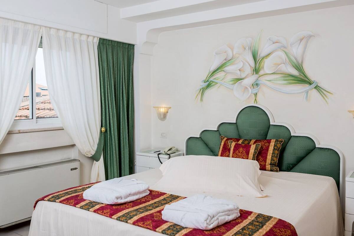 Habitación de hotel con cama, cabecera verde, una obra de arte en la pared con estilo caligráfico, ventana con cortinas y mesita de noche con lámpara.