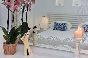 Quarto com cama, mesa de cabeceira, abajur, almofadas decorativas, espelho e uma vela acesa.