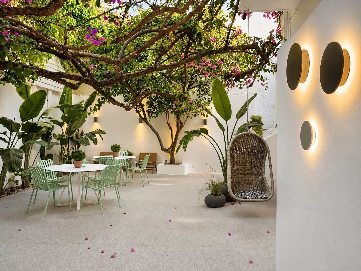An indoor patio with round tables and chairs, surrounded by large green plants and a tree with pink flowers.