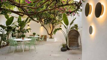 An indoor patio with round tables and chairs, surrounded by large green plants and a tree with pink flowers.