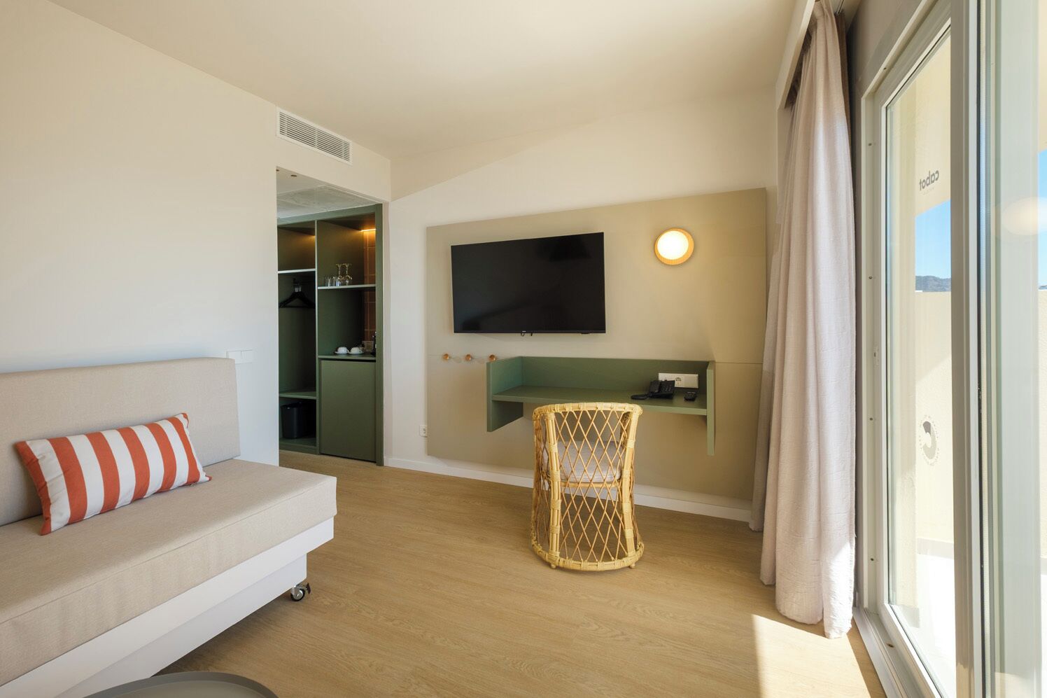Ein modernes Hotelzimmer mit einer Couch, einem Fernseher, einem Korbsessel und Blick durch ein Fenster.