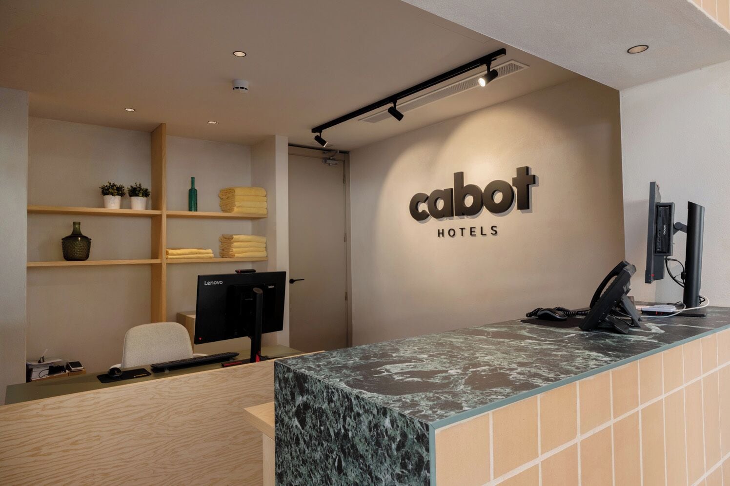 Un mostrador de recepción de hotel con encimera de mármol, un monitor de computadora, un teléfono y estantes con objetos decorativos.