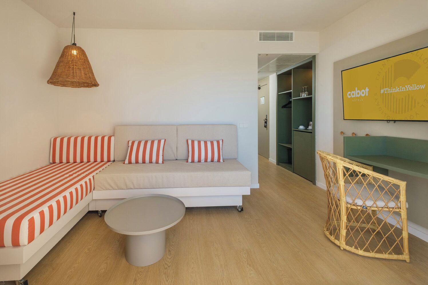 A modern hotel room with a sofa, a small round table, a wicker chair, and a television mounted on the wall.