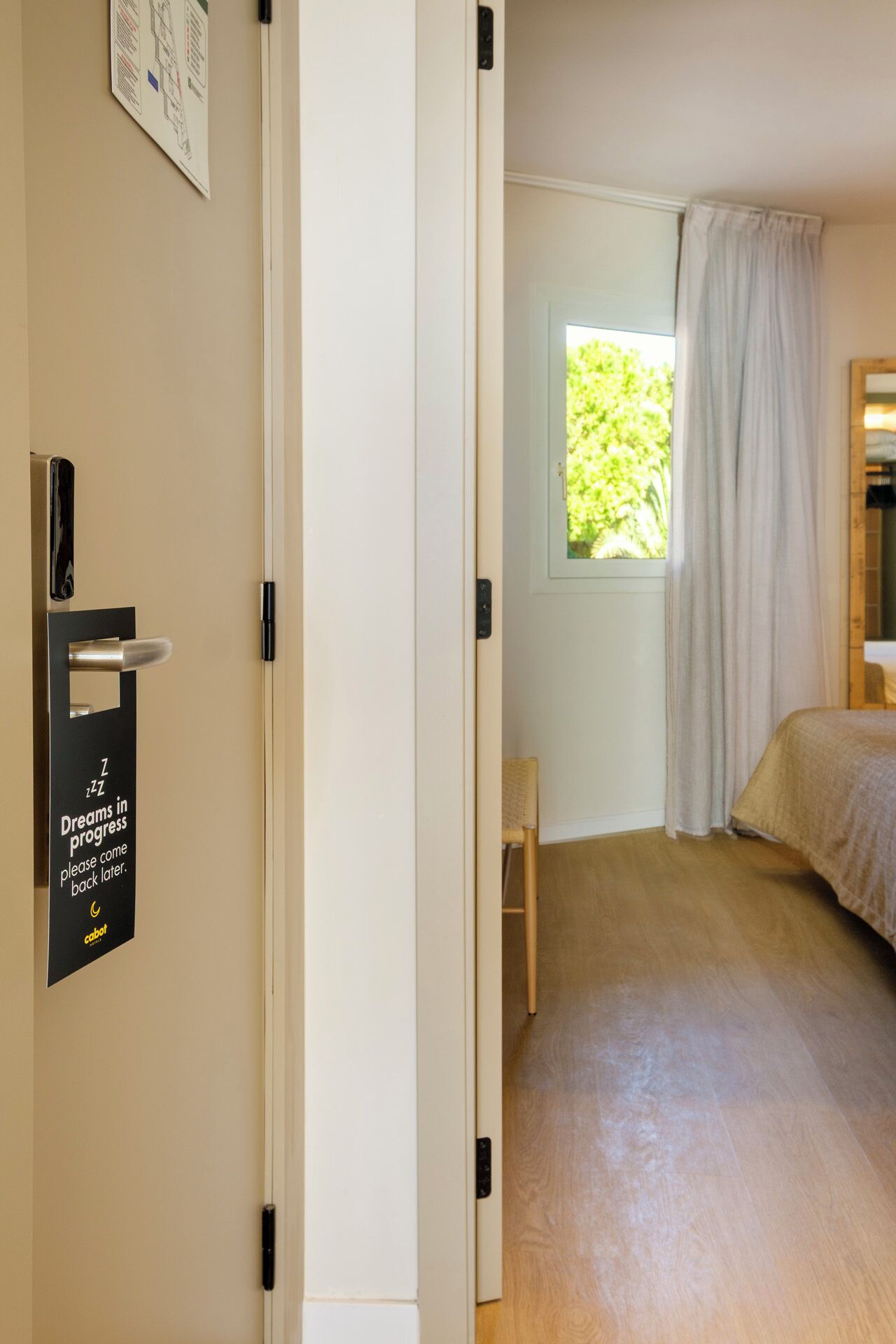 Habitación de hotel con cama, una silla, ventana con cortinas, puerta con un letrero y un cuadro colgado en la pared.
