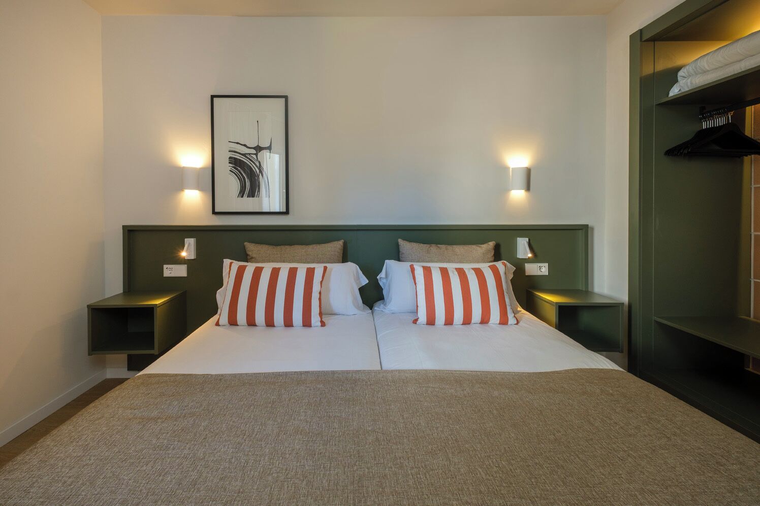 Una cama doble con almohadillas a rayas, dos mesitas de noche con lámparas, una obra de arte abstracta enmarcadas en la pared y un cabecero verde.