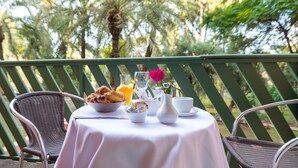 Une table dressée avec des aliments pour le petit-déjeuner, une rose et un verre de jus d’orange, entourée de chaises en osier sur un balcon d’où l’on peut admirer des palmiers.