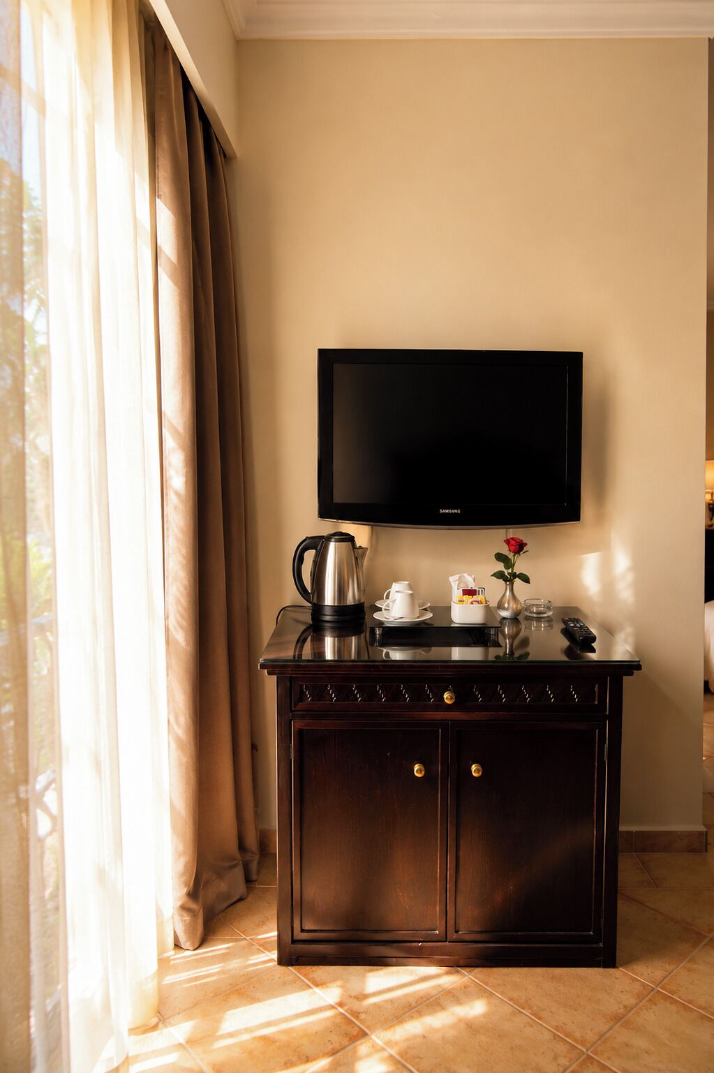 Habitación de hotel con televisión de pantalla plana montada en la pared, un mueble de madera oscura con cajones, una cafetera, una tetera y un florero con una rosa roja.