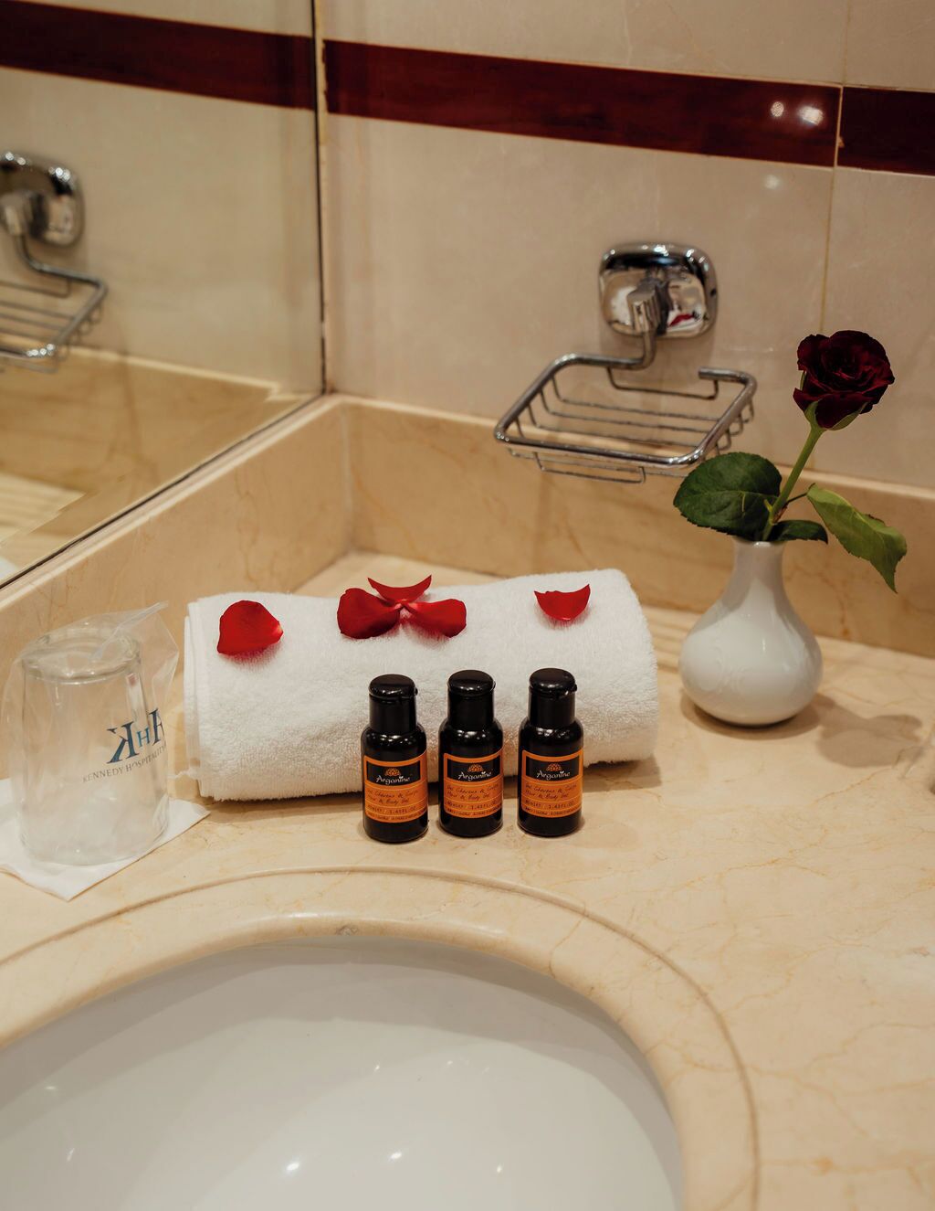 A bathroom with a white towel, rose petals, and three bottles of lotion on the counter.