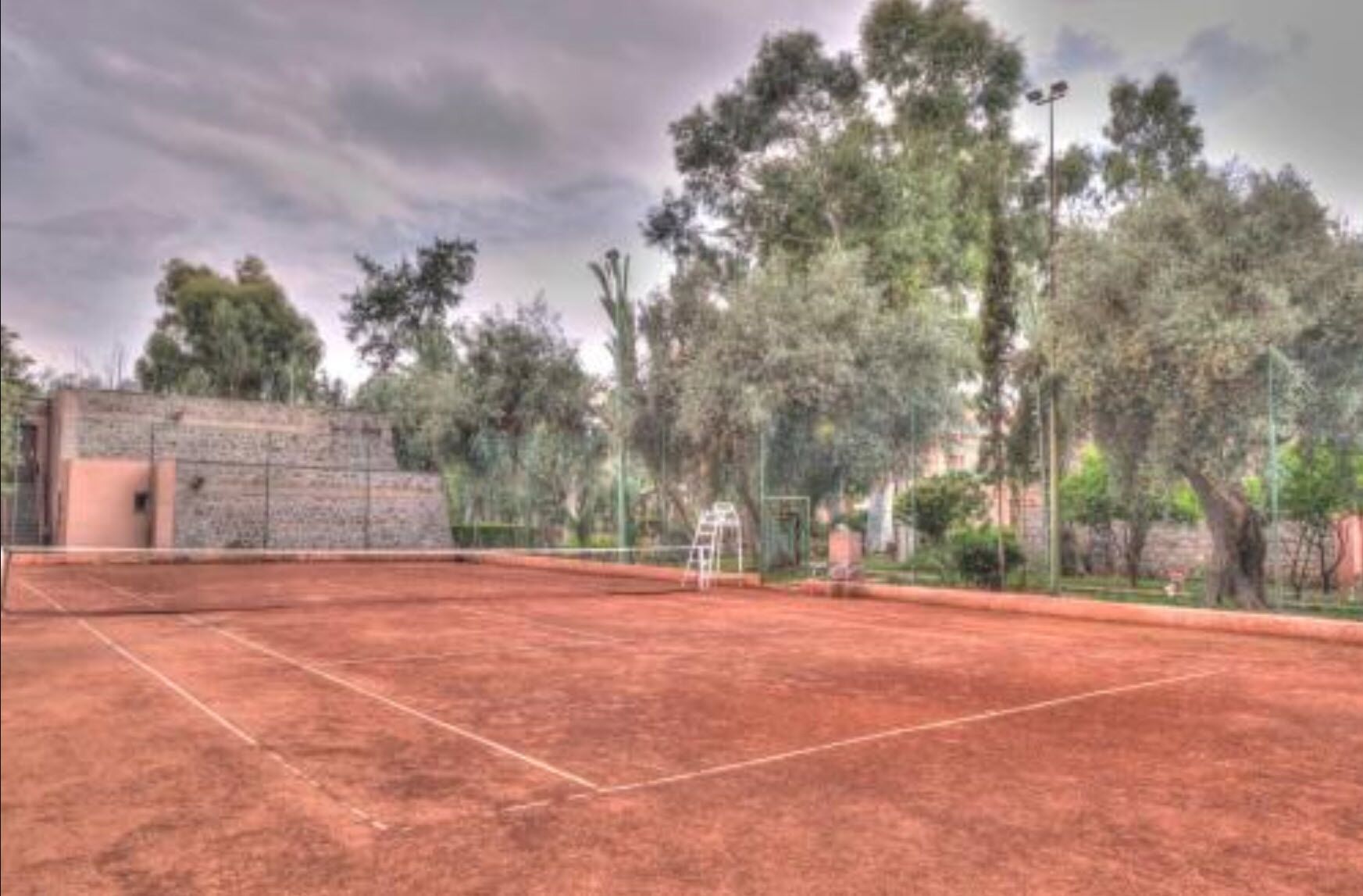 An outdoor tennis court with a brick surface, surrounded by trees and a stone wall.