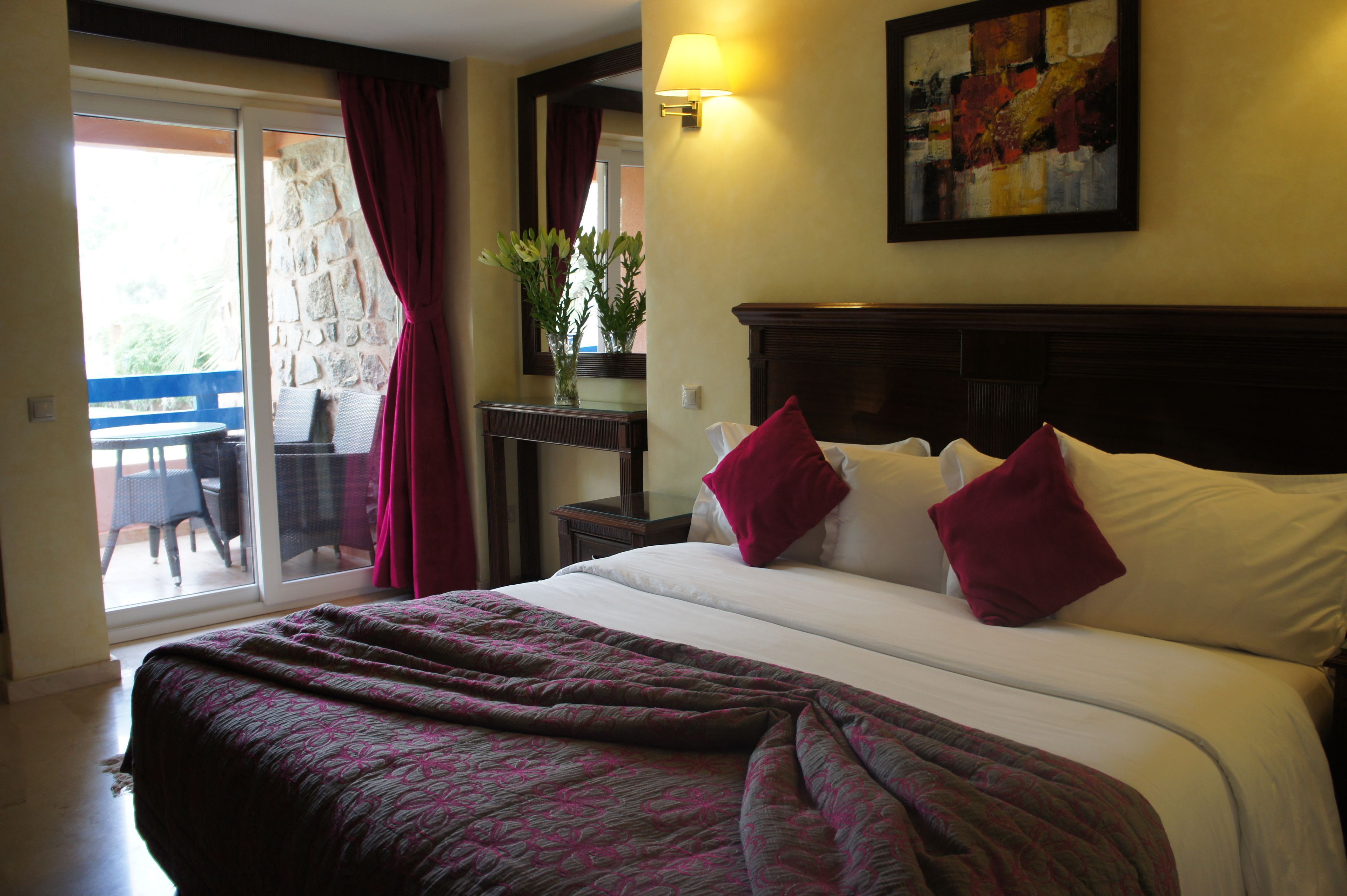 Habitación de hotel con cama, mesita de noche, lámpara, un cuadro y vista al exterior.