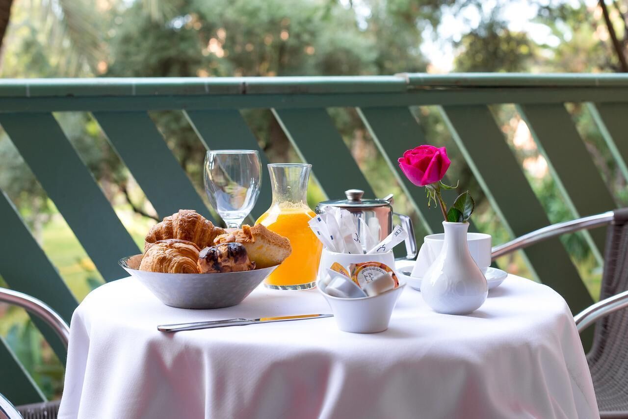 Una mesa con cruasanes, zumo de naranja, una cafetera y un florero con una rosa en un balcón con baranda verde.
