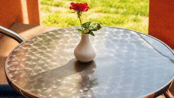 Mesa de vidrio redonda con un florero blanco que contiene una sola rosa roja, ubicada en un patio exterior con vistas a un césped verde y un muro de ladrillo.