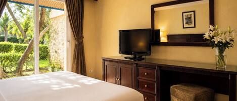 A hotel room with a bed, a desk with a television, a vase with flowers, and a view of greenery outside.