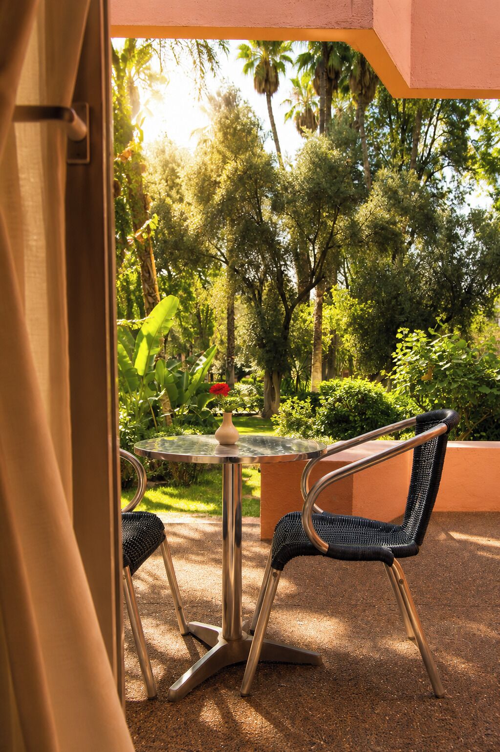 An outdoor seating area with a glass table and two chairs, surrounded by greenery and a view of trees.