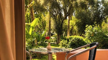 An outdoor seating area with a glass table and two chairs, surrounded by greenery and a view of trees.