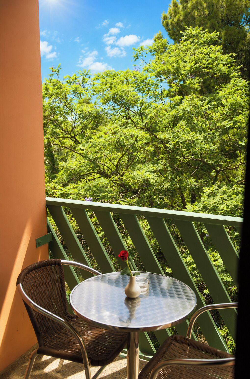 Un balcón con una mesa y sillas, un florero con una rosa roja y vistas a un follaje verde frondoso bajo un cielo despejado.