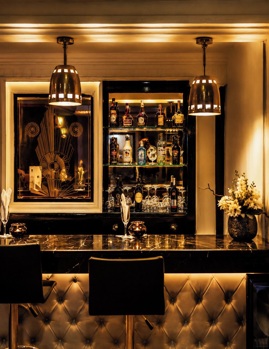 A bar area with a marble countertop, two pendant lights, a selection of liquor bottles, glasses, and a decorative mirror.