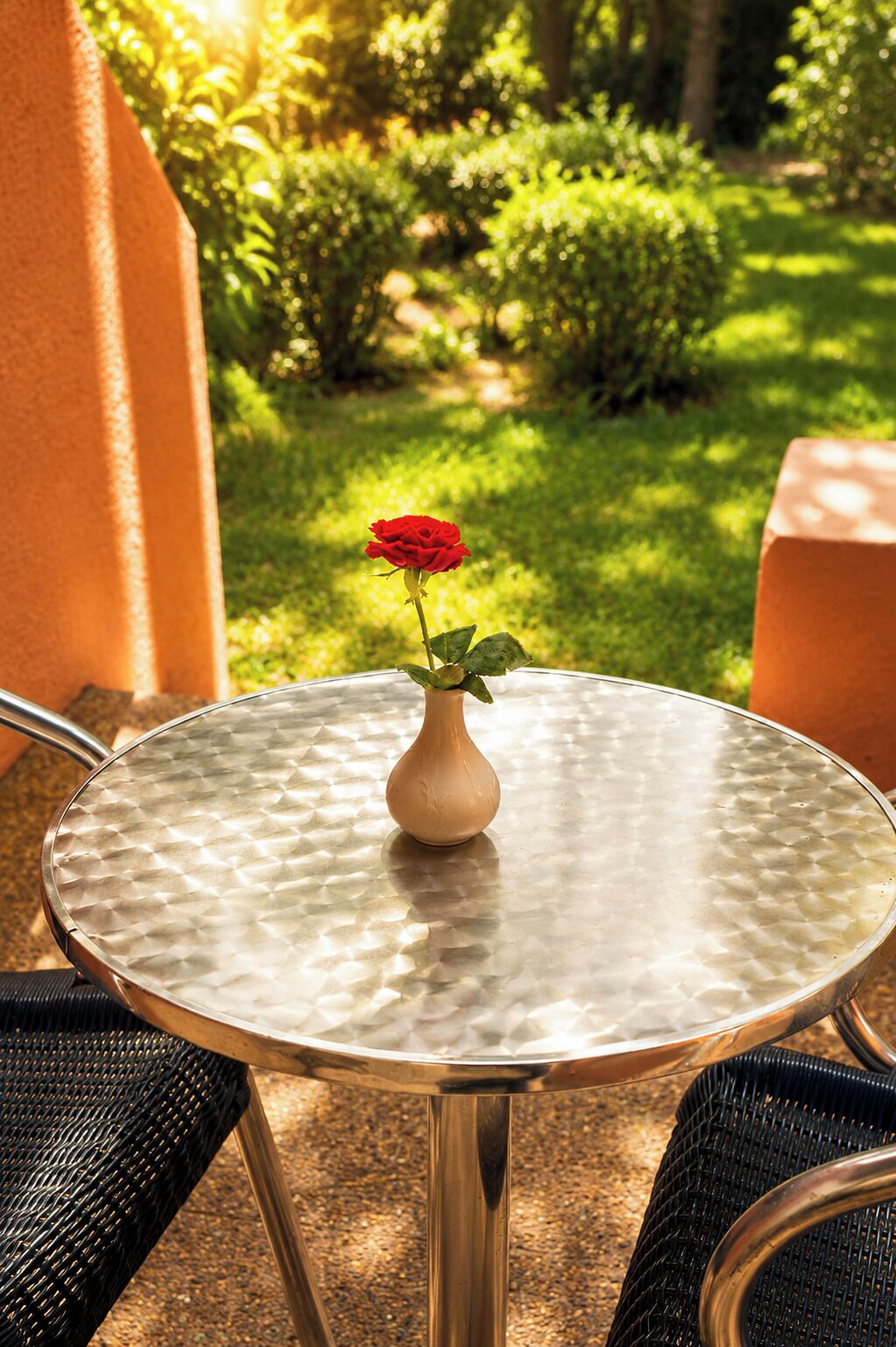 Une table ronde avec un plateau en verre texturé, un petit vase contenant une seule rose rouge et une chaise en osier.