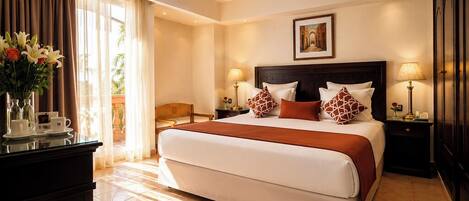 A hotel room with a large bed, two bedside tables with lamps, a view of the outdoors, and a vase of flowers on a dresser.