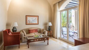 Una sala de estar con un sofá, sillones, una mesa de centro y una mesa de comedor. Hay un ventanal con cortinas, un cuadro en la pared y una lámpara sobre una mesita.