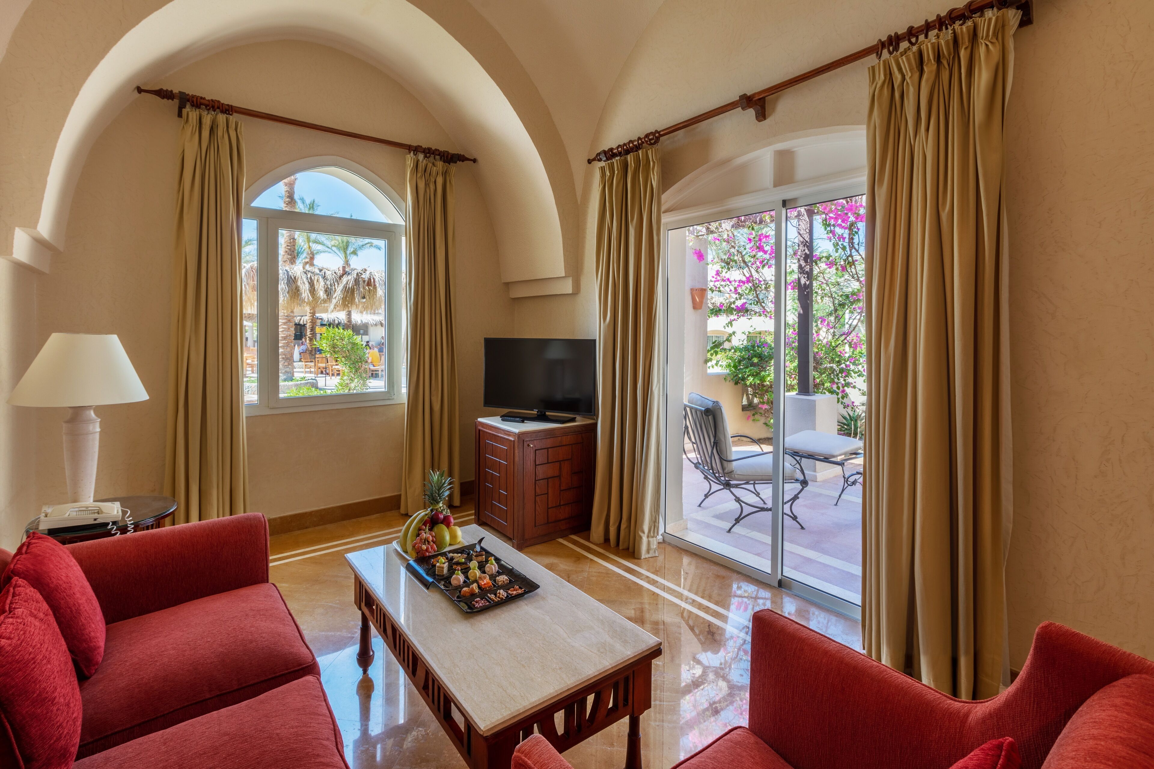 A hotel room with a red sofa, a wooden table, a TV, and a balcony with a chair.