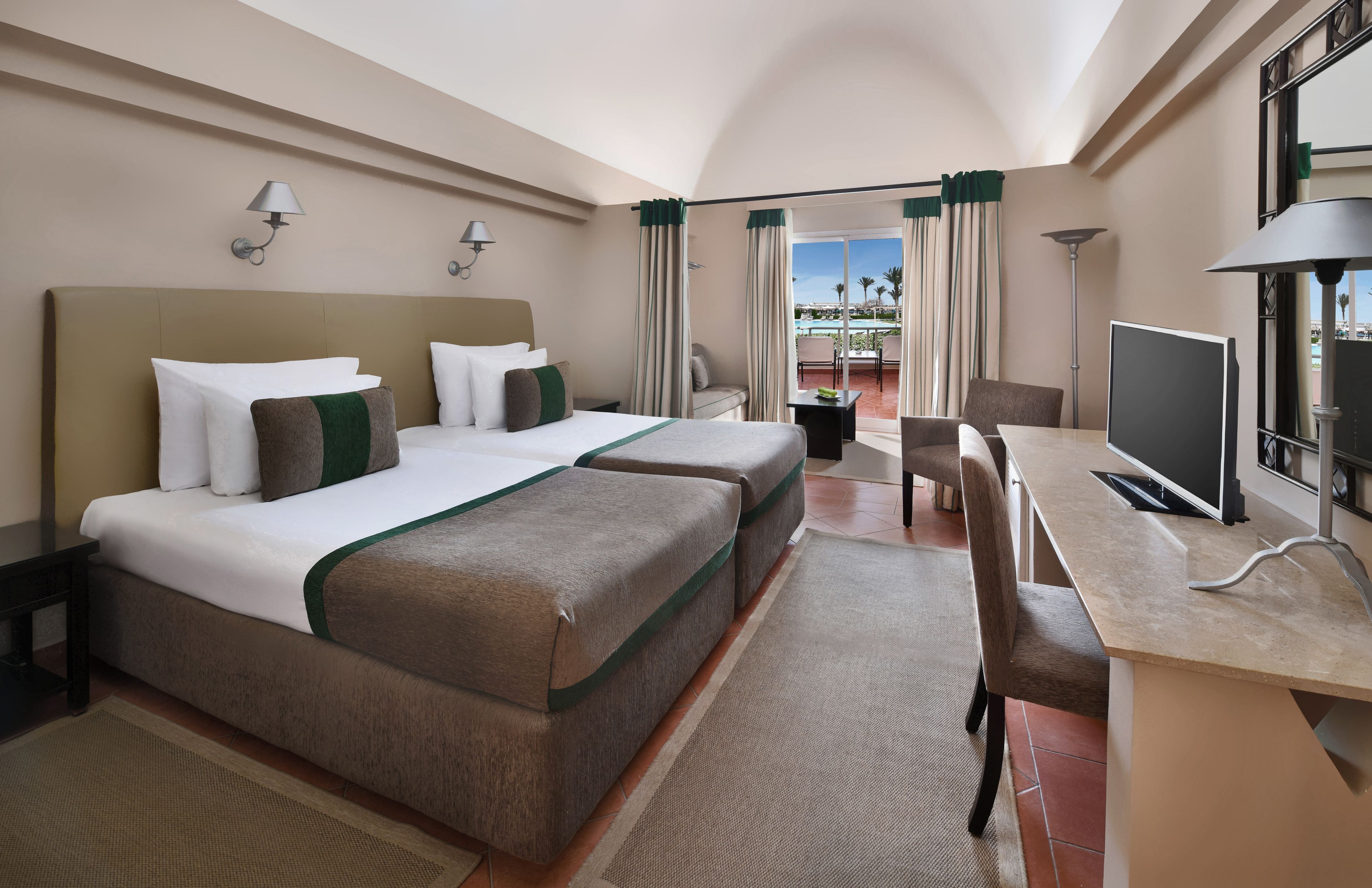 A hotel room with a bed, two bedside tables, a desk with a chair, a TV, and a view of a pool and palm trees.