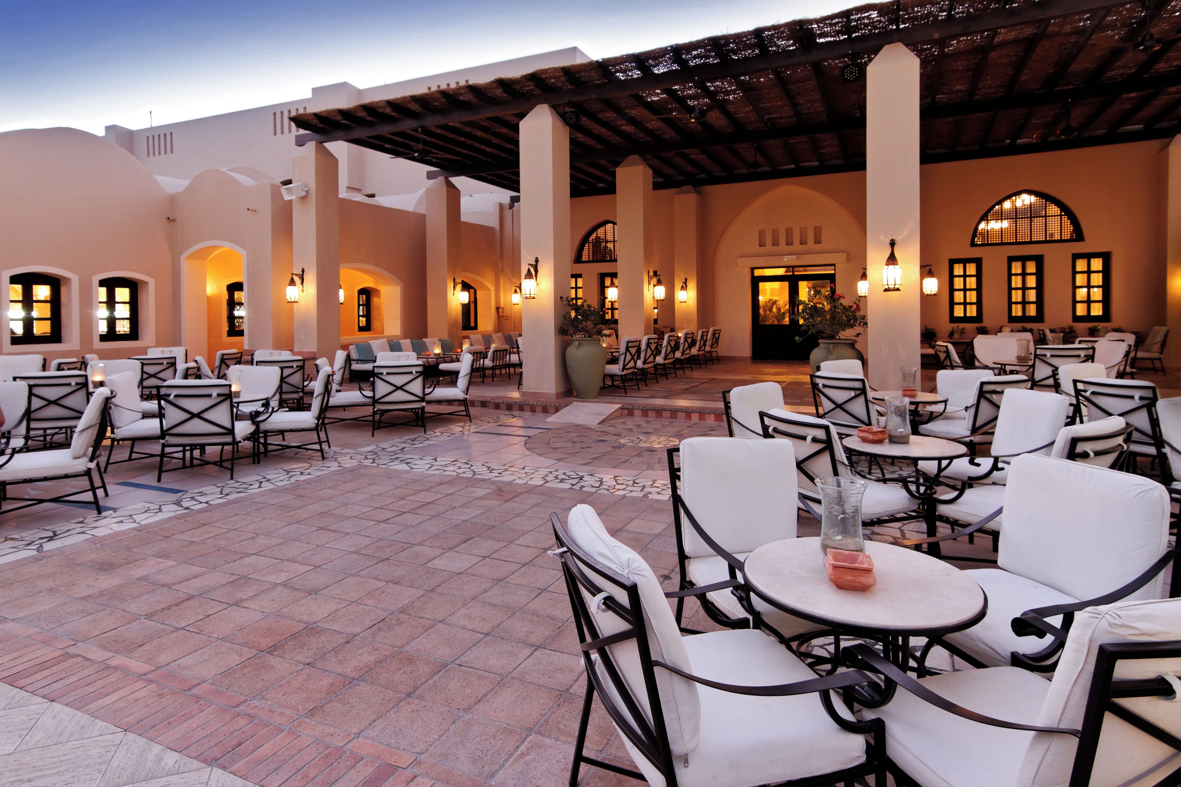 An outdoor patio area with multiple tables and chairs, a tiled floor, and a building with arched windows and columns.