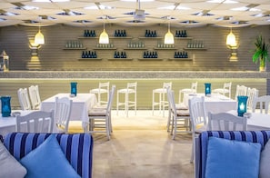 Un restaurante con mesas y sillas blancas, cojines azules y una pared decorada con botellas y vasos.
