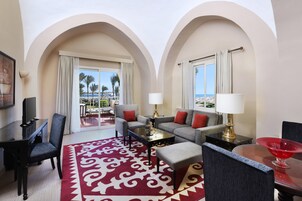 Habitación de hotel con balcón, sofá, mesa de centro, televisor y un cuenco decorativo rojo.