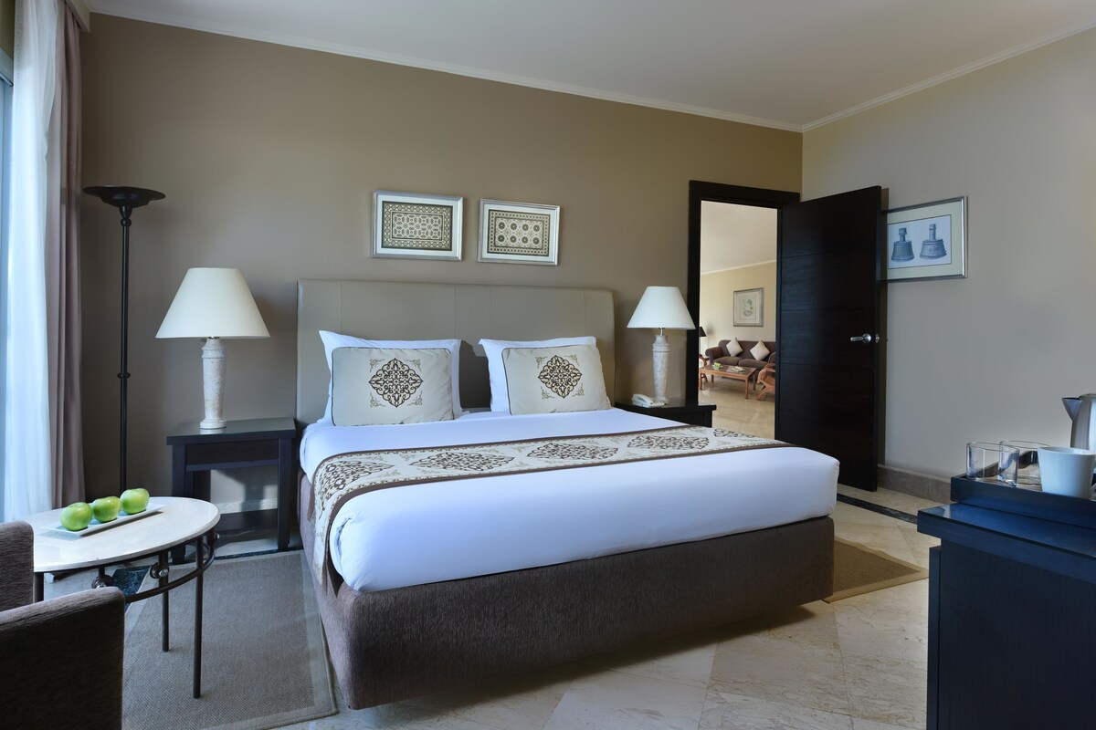 A hotel room with a bed, bedside tables, a desk, a chair, and a view of the adjacent living area.
