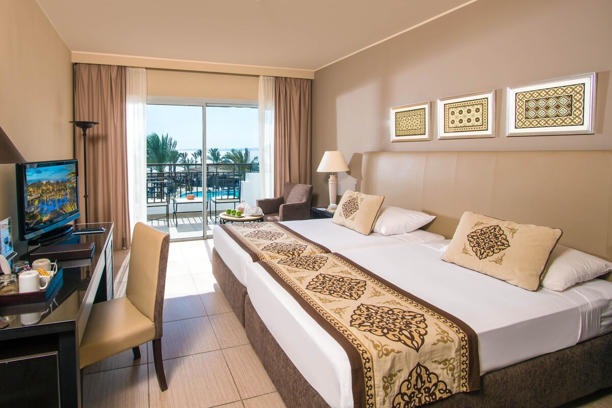 A hotel room with a bed, desk, chair, and a balcony with a pool view.