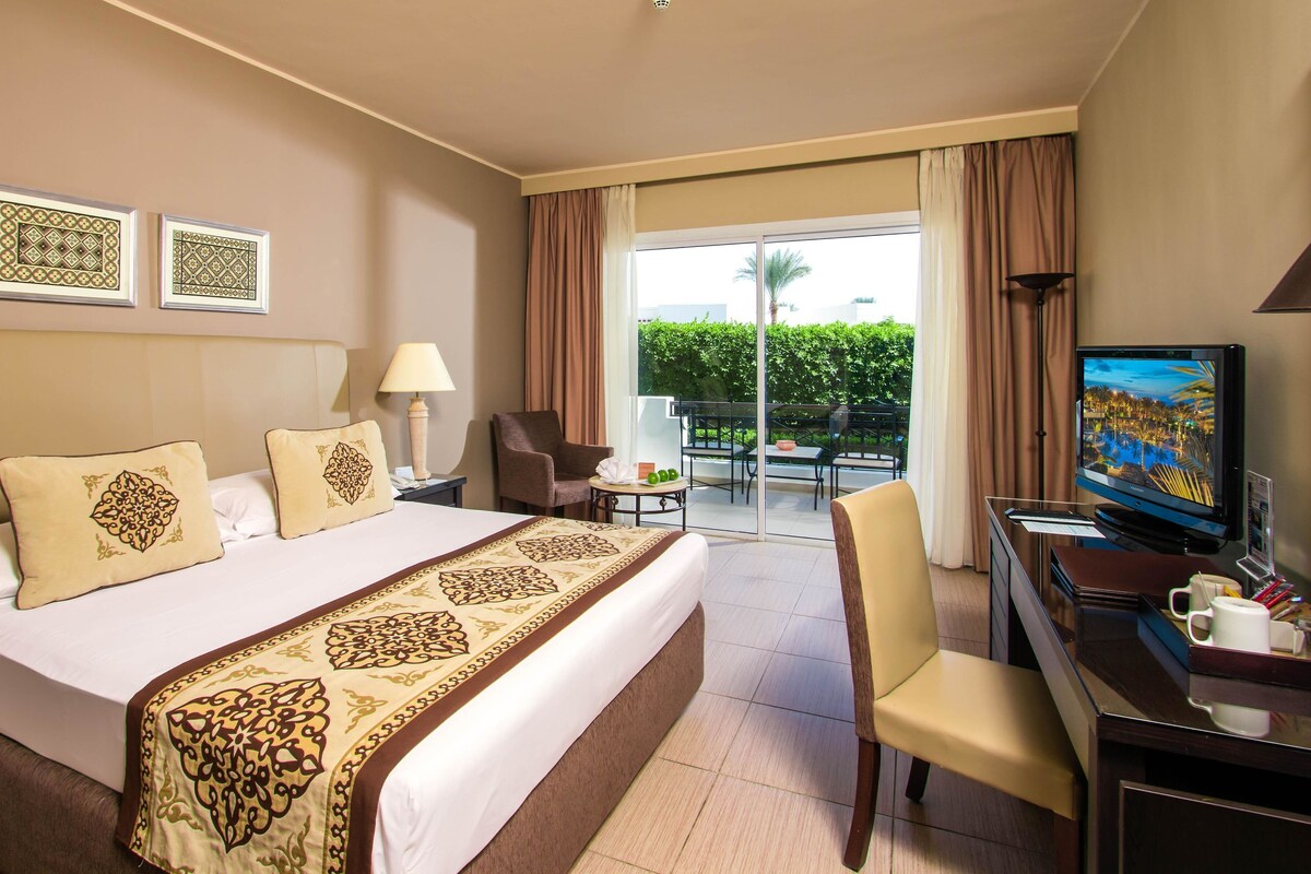 A hotel room with a large bed, a desk with a TV, a chair, and a balcony with a view of greenery.
